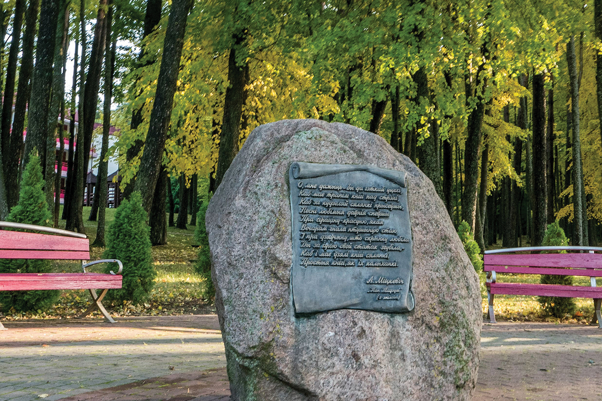 Głaz pamiątkowy z elementem dekoracyjnym w postaci metalowego zwoju z fragmentem z „Pana Tadeusza” Adama Mickiewicza. Po obu stronach pomnika stoją ławki przy chodniku. Za nimi zielony las z prześwitującymi w tle zabudowaniami. 